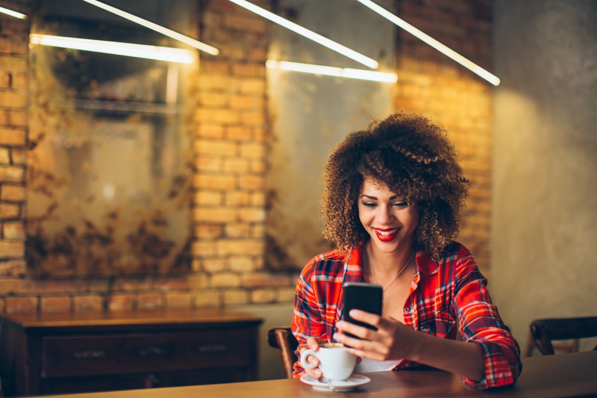 young black woman in plaid shirt drinking coffee and smiling at her cell phone