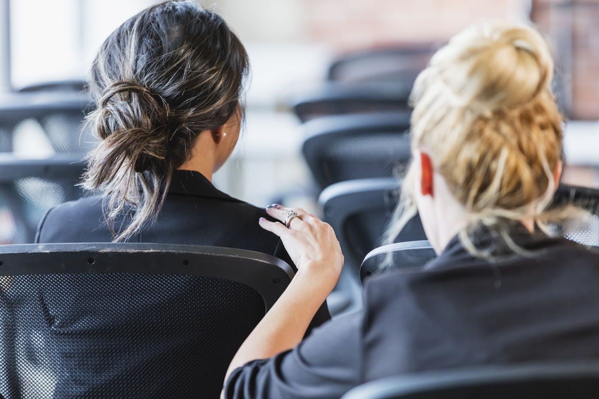 The back of the heads of two businesswomen sitting in chairs, attending a business conference or seminar. One woman is tapping the other on the shoulder trying to get her attention.