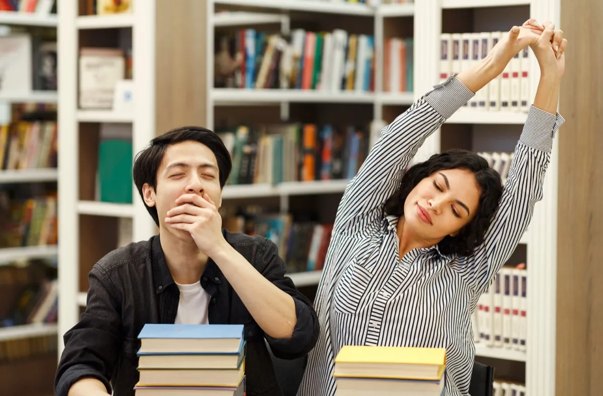 Girl stretching boy yawning studying in the library with books