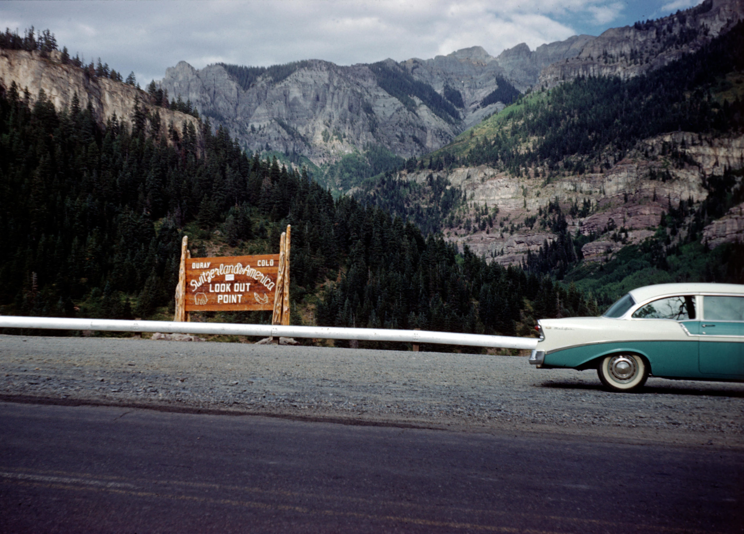 a turquoise car drives on a mountain side road in 1960