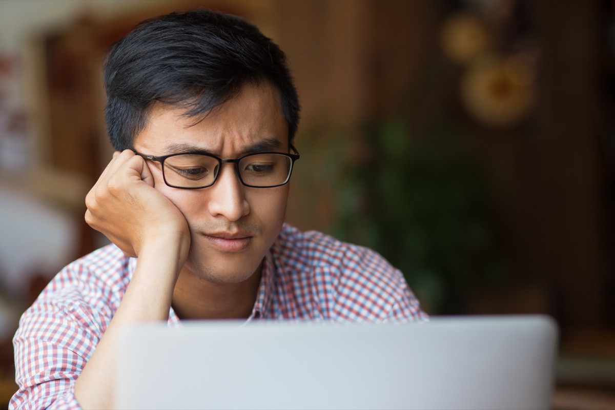 Portrait of bored young male student wearing glasses sitting at laptop leaning on arm