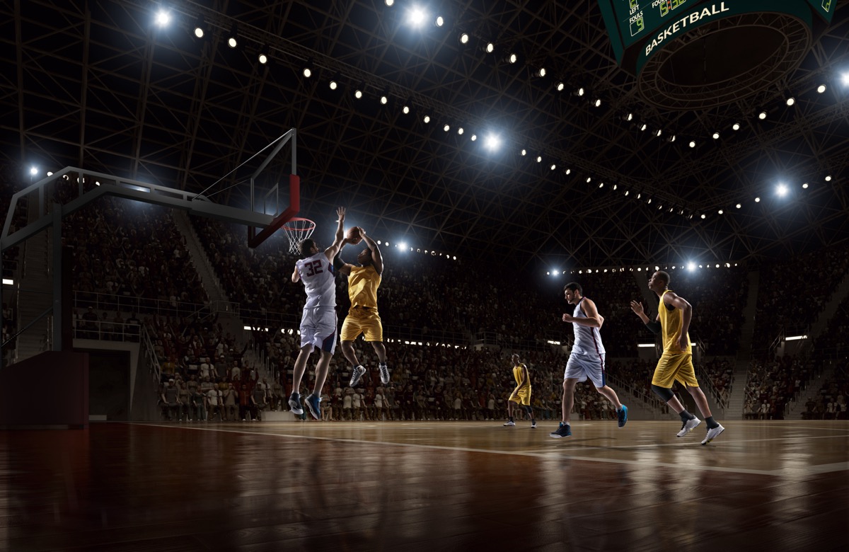 Low angle view of a professional basketball game. A player is in mid air holding ball about to score a slam dunk, but the player from the opposite team is ready to block him. A game is in a indoor floodlit basketball arena. All players are wearing generic unbranded basketball uniform.