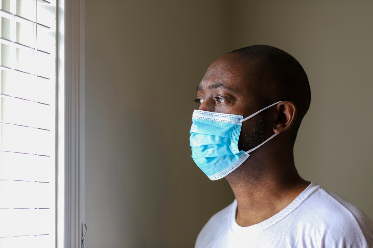 An African-American man wearing a protective face mask to prevent virus infection while staring out window