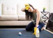 Young woman rubbing head wearing casuals and yellow gloves cleaning carpet with liquid soap