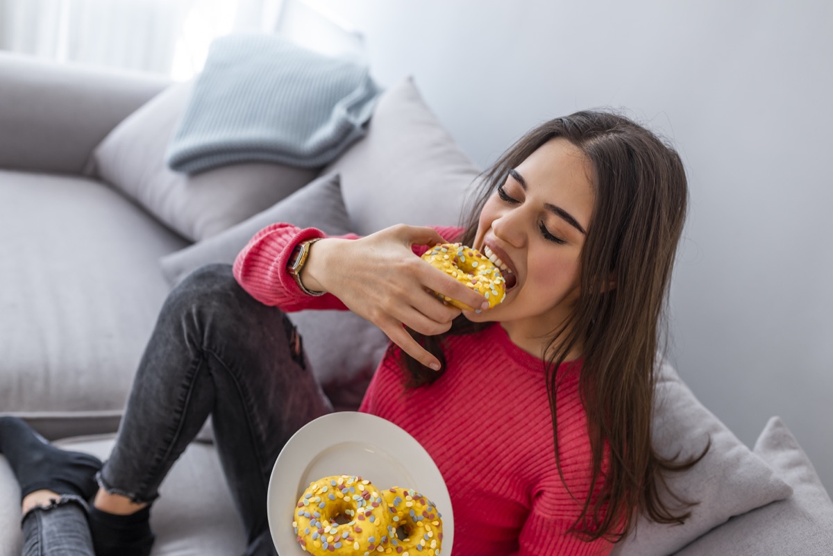 Woman is eating tasty sugary donuts. Portrait of beautiful young woman eating donuts at home. Woman sitting on the sofa while eating a plate of donuts
