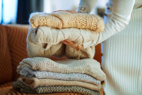 Closeup on stylish woman in white sweater and skirt in the modern living room in sunny winter day near couch folding sweaters for storage.