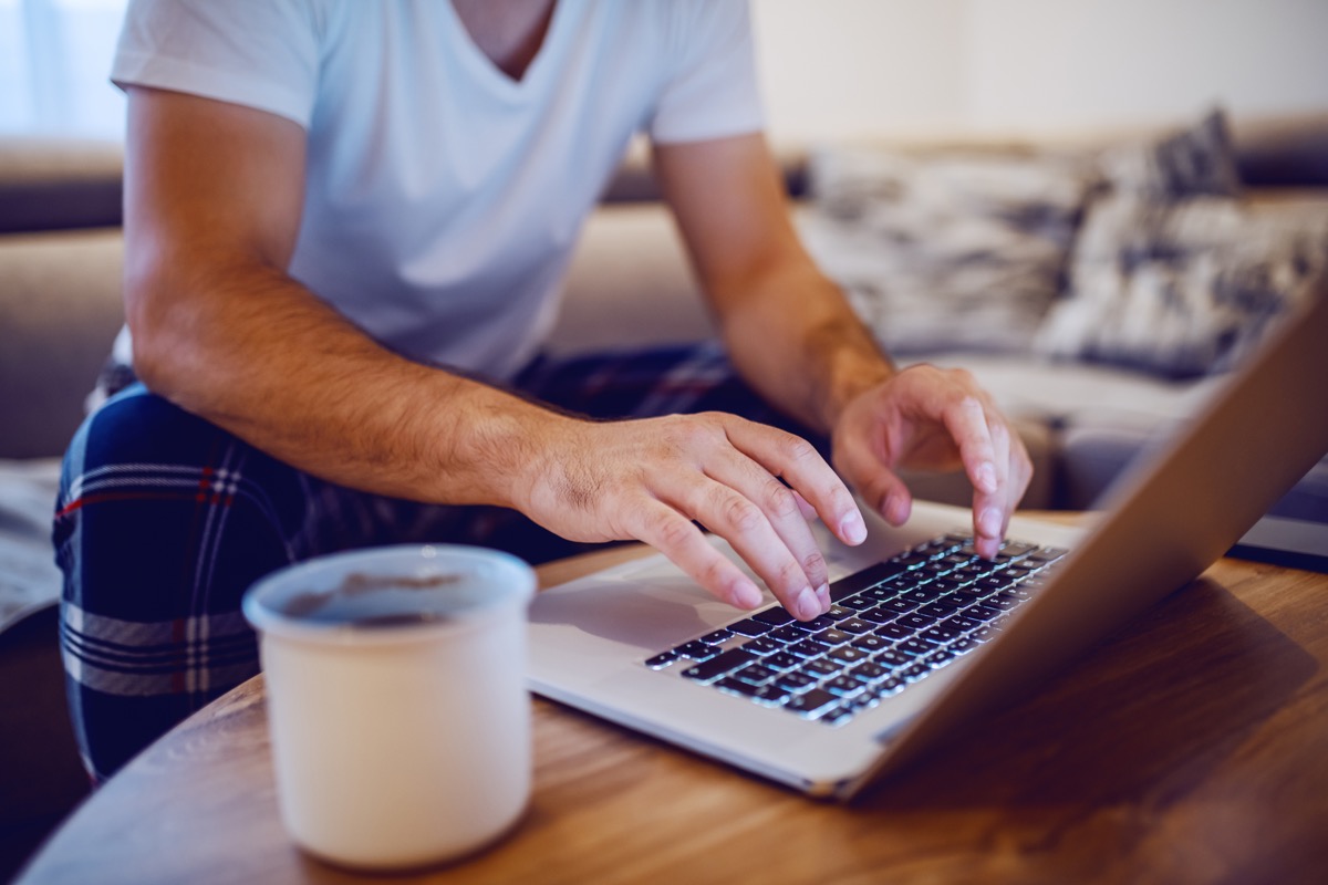 Cropped photo of man in pajamas typing on laptop keyboard and surfing the internet while sitting on sofa in living room in morning. Selective focus on hands.