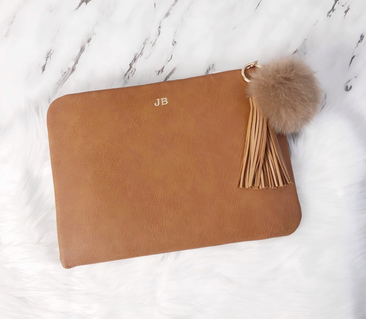 brown leather zip-top case with gold embroidered initials, a gray pom pom, and a brown tassel