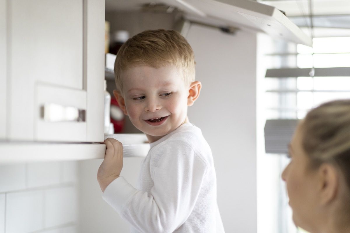 A shot of a mother and her son in the kitchen, the young boy is reaching in the cupboard.