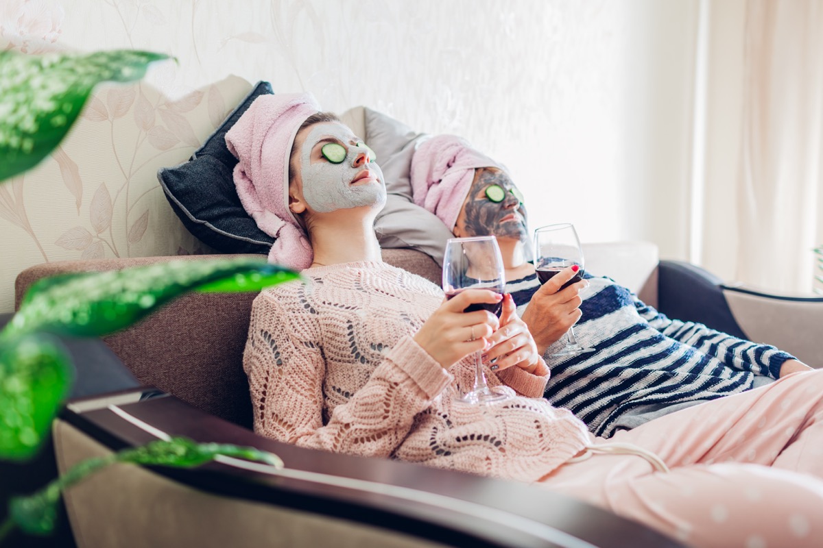 Mother and her adult daughter applied facial masks and cucumbers on eyes. Women chilling while having wine sitting on couch at home