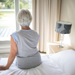 Rear view of senior woman sitting on bed in bedroom, looking out window