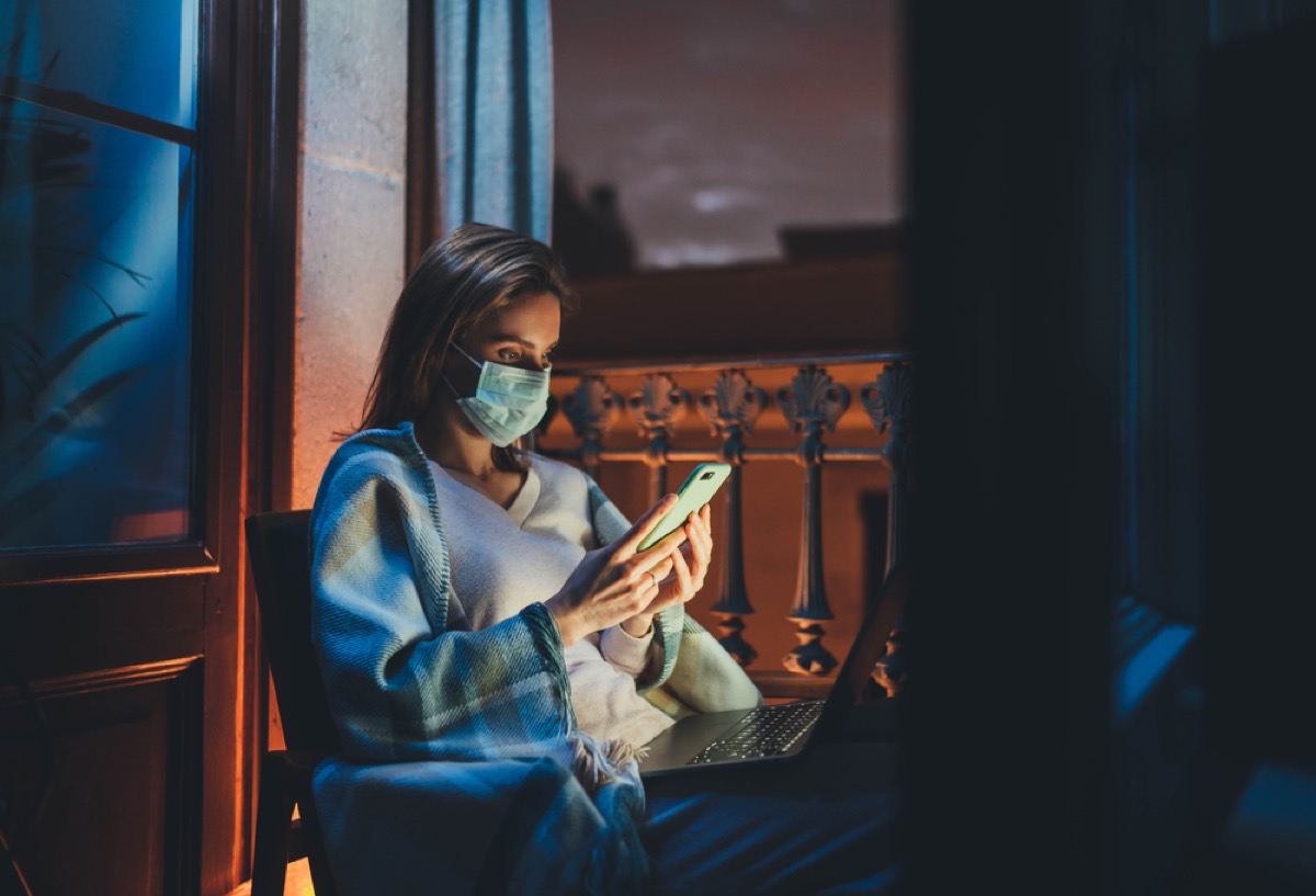 woman with a face mask sitting at a window looking at her phone and computer