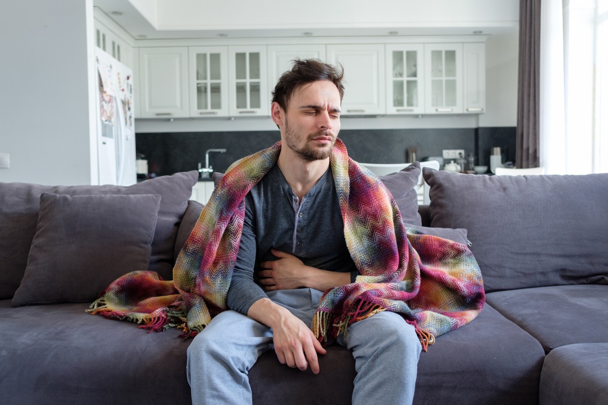 A young sick white man sits on a sofa in blankets and holds his stomach