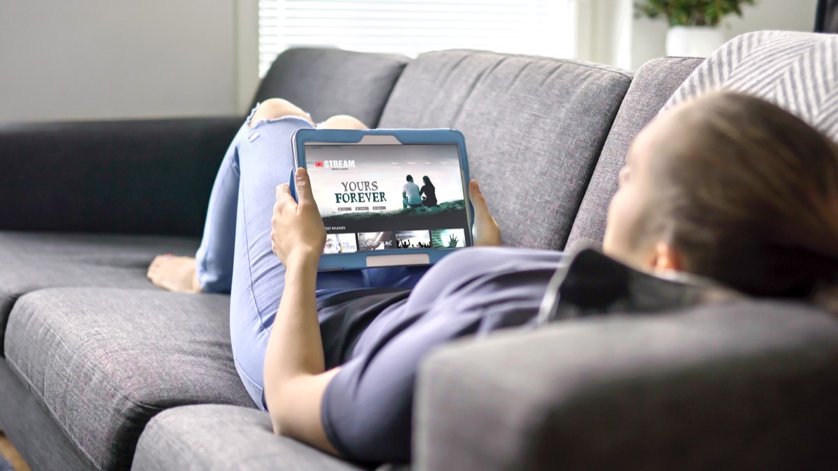 O femeie selectează un film dintr-un serviciu de streaming online folosind o tabletă.  Urmărește seriale cu conceptul de site video la cerere (VOD).  Redați filmul digital de pe site prin Network TV.  Machetă pe ecranul unui dispozitiv inteligent.