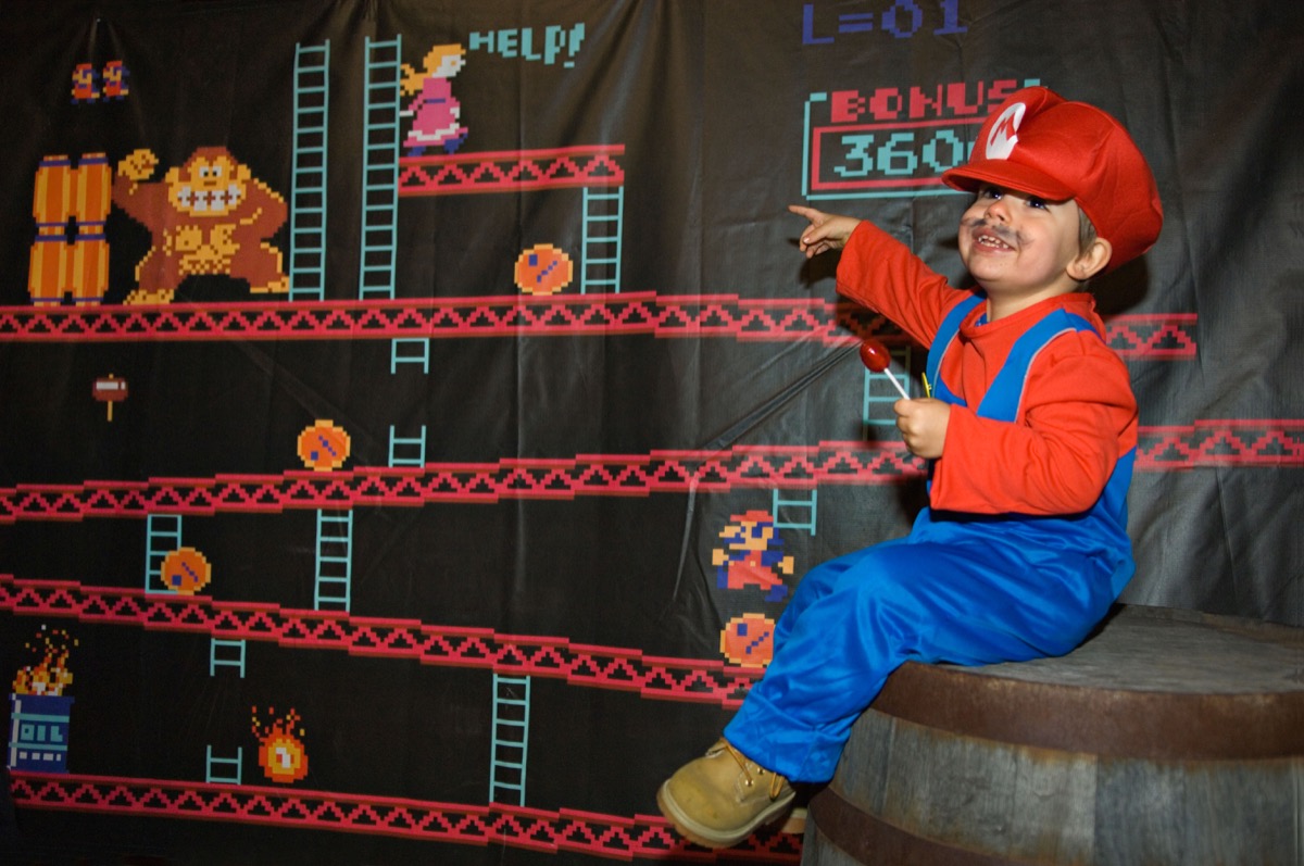 DJ74EX Toddler Dressed Up as Mario Sitting on Barrel with Donkey Kong Banner Behind at the Louisville Zoo Halloween Party in Louisville
