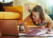 smiling healthy woman in fitness clothes in the modern living room watching fitness tutorial on internet via laptop and doing pushups on fitness mat. (smiling healthy woman in fitness clothes in the modern living room watching fitness tutorial on inte