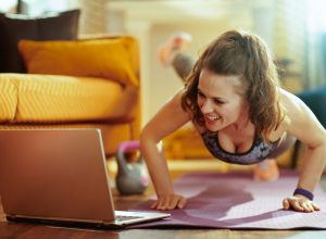 smiling healthy woman in fitness clothes in the modern living room watching fitness tutorial on internet via laptop and doing pushups on fitness mat. (smiling healthy woman in fitness clothes in the modern living room watching fitness tutorial on inte
