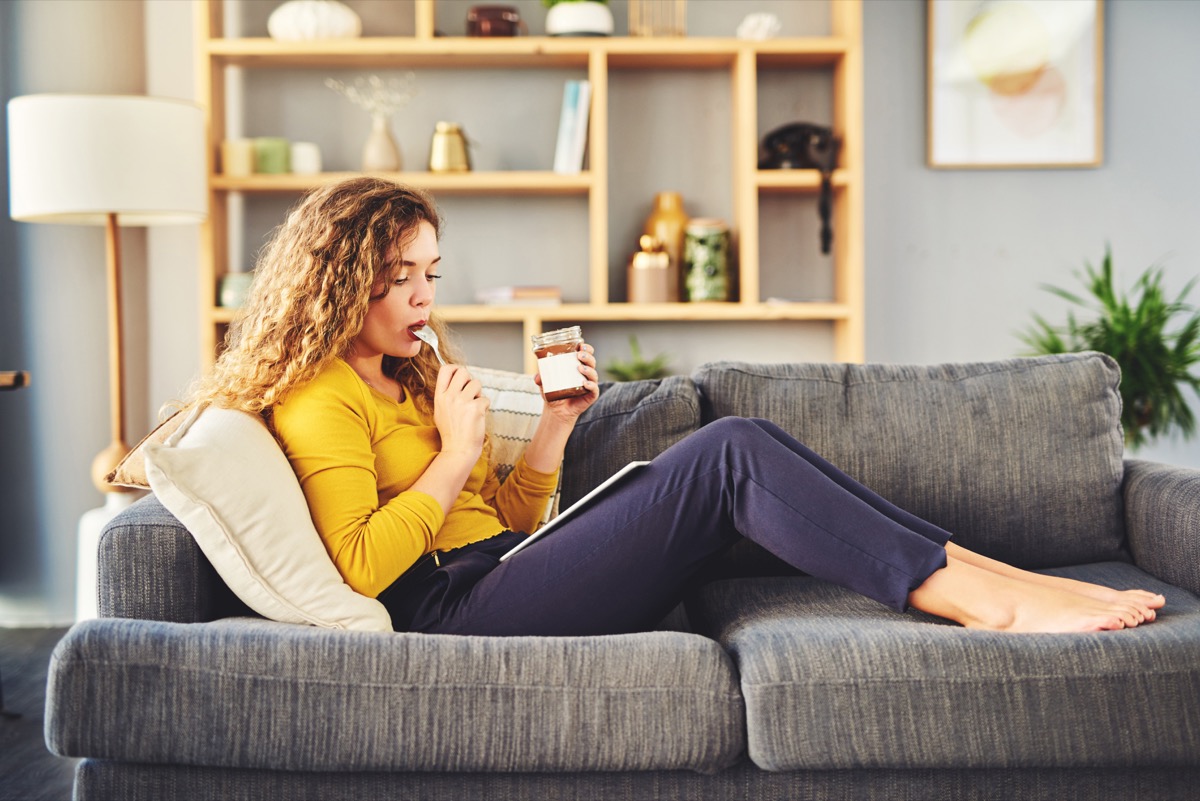 Shot of a young woman eating chocolate from a jar while relaxing on the sofa at home