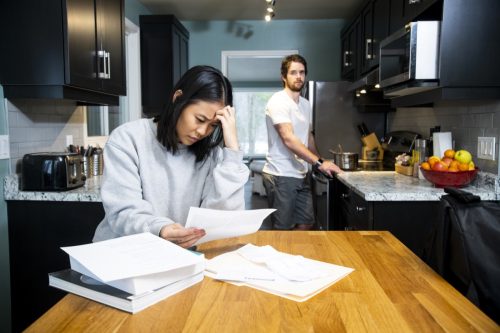 A female student worrying about financial issues at home in her apartment with her male friend cooking at the stove.