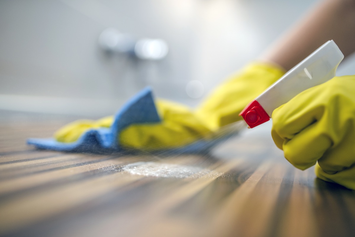 Photo of Woman cleaning kitchen cabinets with sponge and spray cleaner. Female Using Spray Cleaner On Wooden Surface. Maid wiping dust using a spray and a duster while cleaning her house wearing yellow protective gloves, close-up