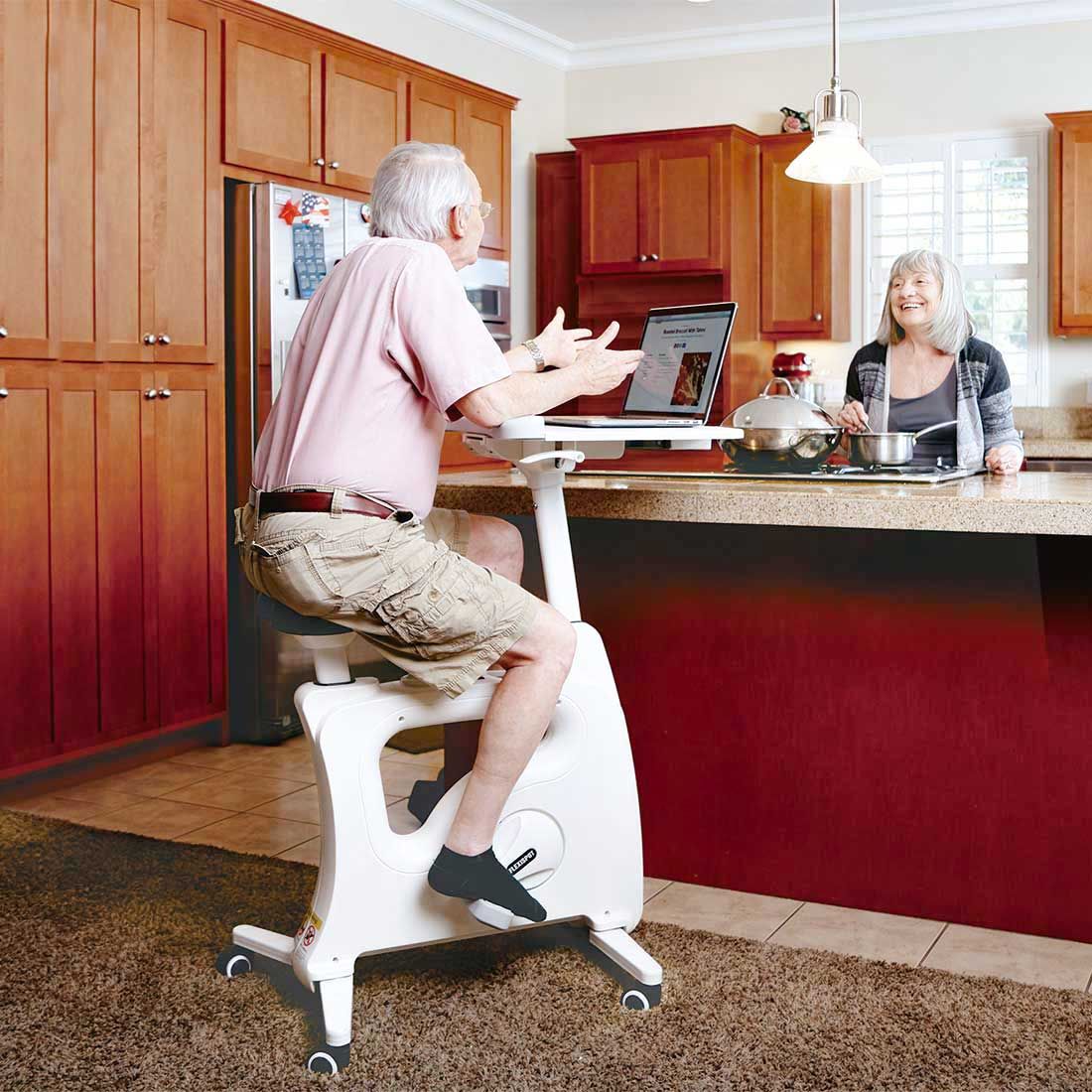 senior man using desk bike in house while talking to wife in kitchen