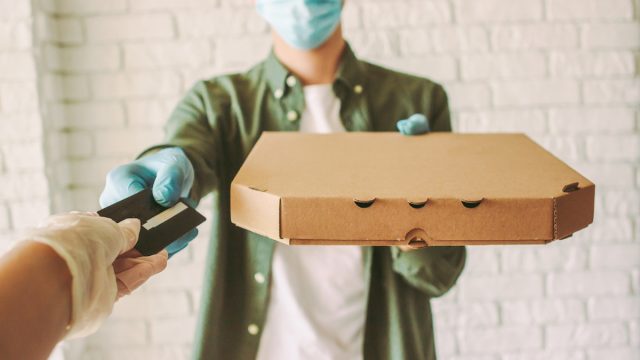Hipster guy delivery service worker in medical face mask and gloves hold pizza box in hand, checkout customer with credit card