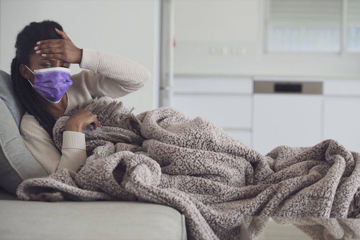 Young ill woman holding hand on forehead, checking temperature, resting, lying on the couch with a cozy blanket. Using purple face mask to prevent other people from getting infected.
