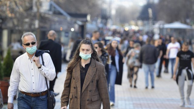 A young woman and an older man are walking on street wearing face masks while others are not