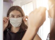 young girl wearing medical protective face mask looking mirror stay at home social dictancing concept