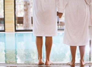 Rear view of man and woman standing on poolside barefoot, holding hands. They are wearing white bathrobes.
