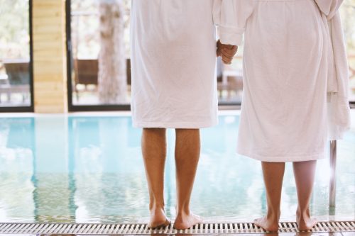 Rückansicht von Mann und Frau, die am Pool stehen, barfuß, Händchen haltend.  Sie tragen weiße Bademäntel.