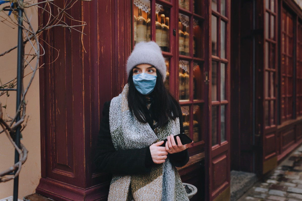 A young girl in a coat, a scarf, a hat and a protective medical blue mask is stand near wooden wall the deserted narrow old street of the city. The concept of quarantine, coronavirus, pandemic COVID-19.