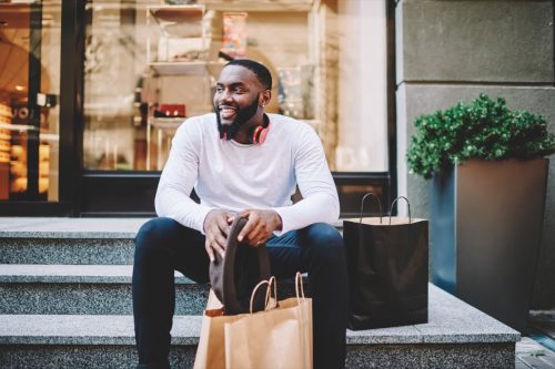 Smiling male customer in trendy wear sitting on stairs of store with bags with copy space for label, cheerful dark skinned hipster guy recreating after shopping and buying purchases
