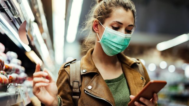 Young woman wearing protective mask on her face and reading shopping list on mobile phone in grocery store during virus pandemic.