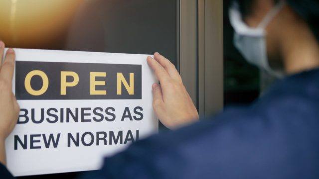A man wearing a mask puts up an open sign that says business as new normal