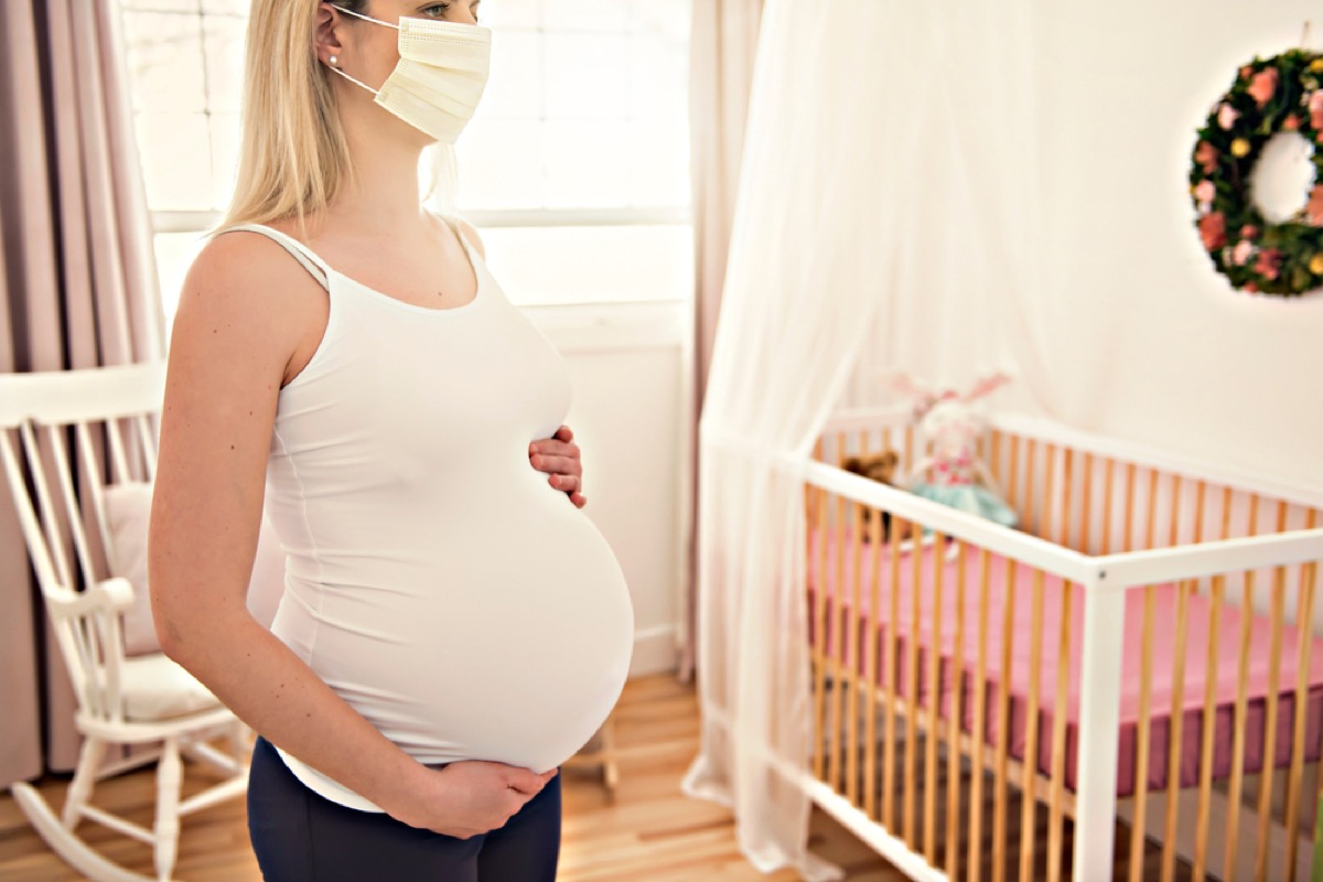 pregnant white woman wearing a face mask and standing by a crib