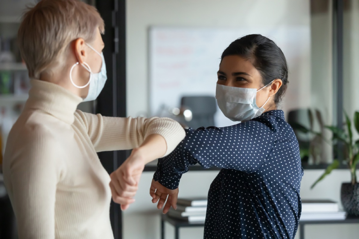 two female office workers wearing face masks greeting each other by touching elbows