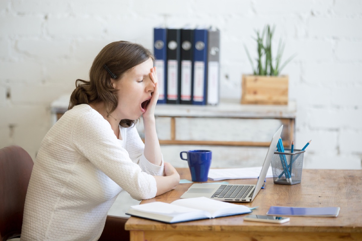 young white woman yawning at her desk in front of a laptop