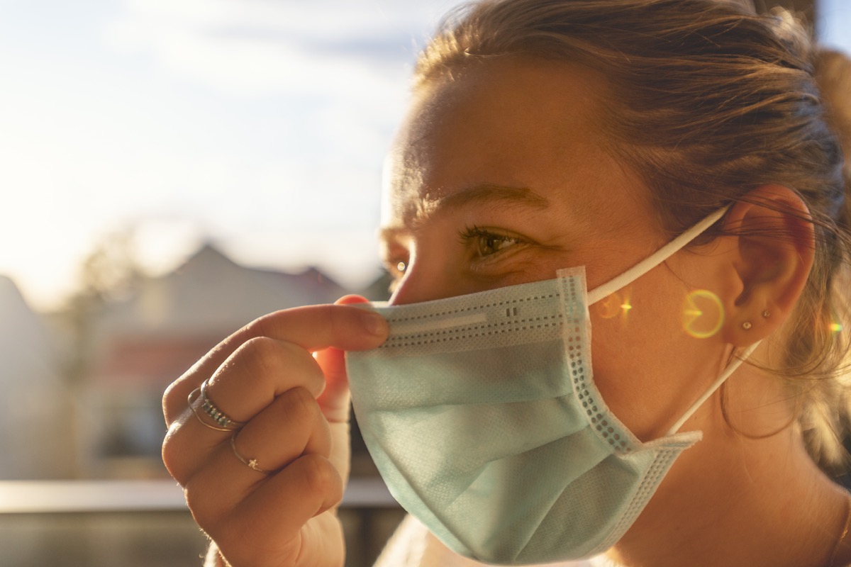 Woman putting on a surgical face mask. She is applying it to her face and is adjusting the mask for a proper fit. Back lit at sunset.