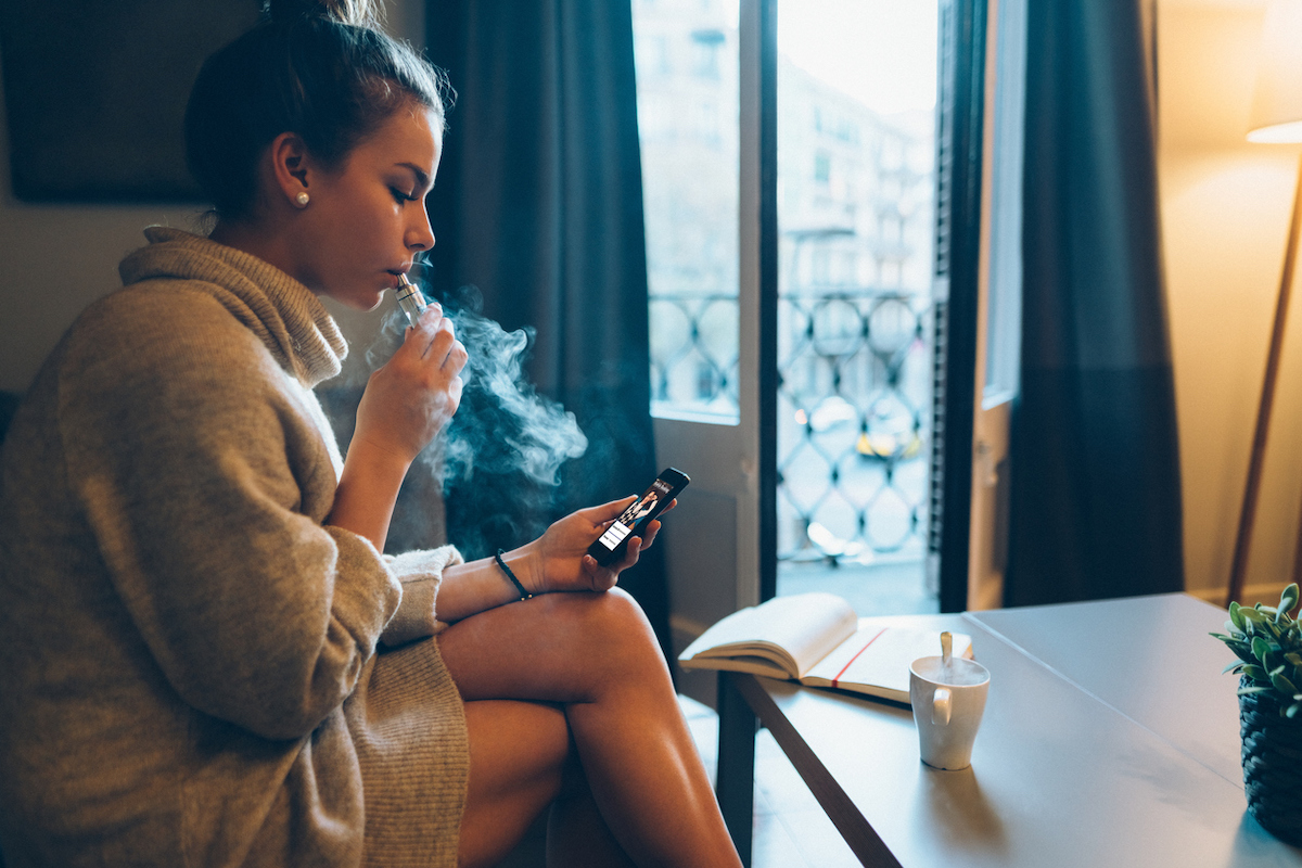 Young woman at home smoking electronic cigarette and spending time on mobile phone