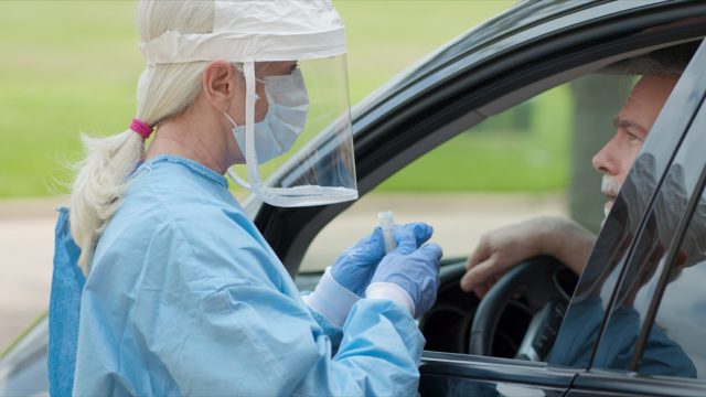 White female nurse in face shield testing mature white man in car for coronavirus