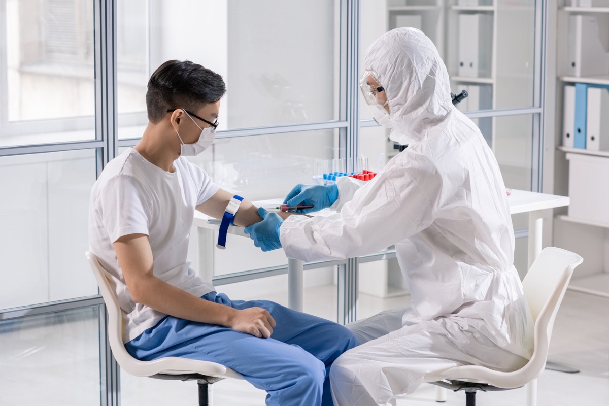 Young man in mask sitting in lab while doctor in protective coveralls taking his blood in syringe for analysis