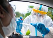 A doctor wearing a face shield and protective suit administers a coronavirus test to a patient sitting in their car.
