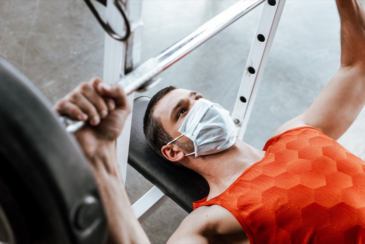 selective focus of sportsman in medical mask exercising with barbell in gym