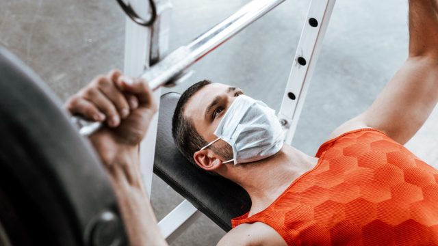man in medical mask exercising with barbell in gym