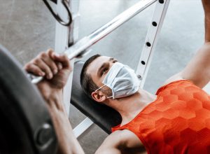 man in medical mask exercising with barbell in gym