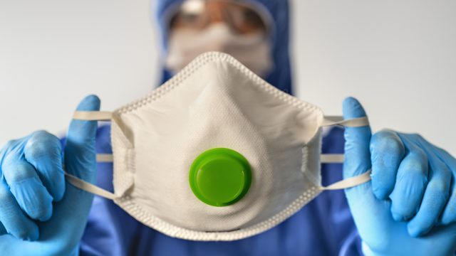 A healthcare worker in full protective gear holds up a face mask with a breathing valve