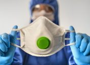 A healthcare worker in full protective gear holds up a face mask with a breathing valve