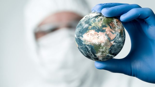 A scientist wearing a face mask and blue gloves holds a small globe in his hand and examines it