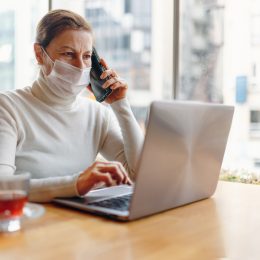 senior Woman With Face Mask, sitting at computer while on the phone
